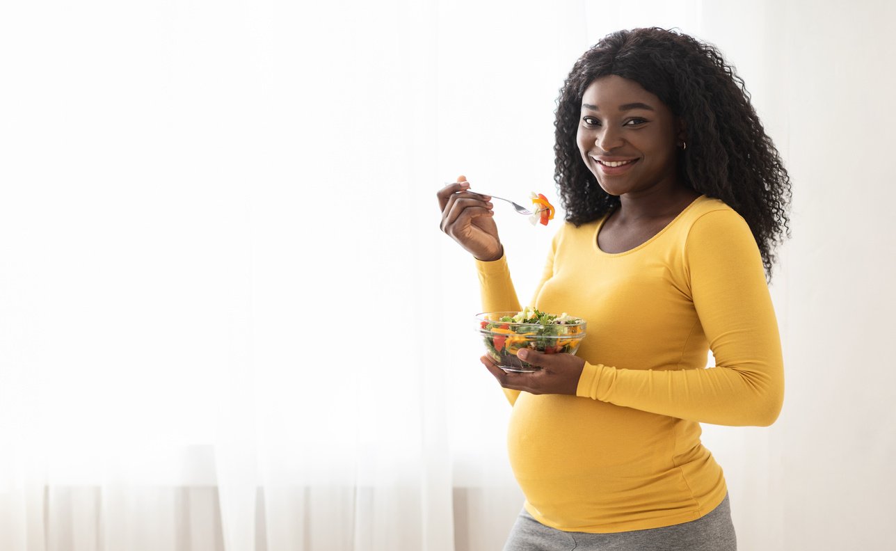 Happy pregnant black woman eating fresh salad at home